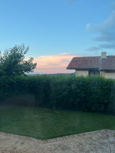 This image, set in Cherni Vrah, Bulgaria, depicts a tranquil residential backyard scene during either sunrise or sunset. The sky, filling the upper portion of the frame, transitions from a clear blue at the left to soft pink and orange hues on the right, indicative of dawn or dusk, with scattered white and grey clouds.
A house with a red-brown terracotta tiled roof and a light-colored chimney is partially visible in the mid-ground, extending from the middle-left towards the center. In front of the house and stretching across the mid-ground is a dense, well-maintained green hedge, which defines the boundary of the property. Behind the hedge, a hazy, darker green expanse suggests a distant forested area or hills. A thin power line is faintly visible traversing the sky, particularly against the colorful clouds.
In the foreground, a vibrant green lawn occupies the lower-right section, appearing freshly mown. Adjacent to the lawn, a textured, light-colored paved area, possibly stone or concrete, forms a patio or walkway. This paving features a distinct, darker-toned border pattern. On the lower-left, the dark green foliage and branches of a tree are prominent, reaching into the frame and silhouetted against the bright sky.
The scene is devoid of any human activity or visible text, conveying a sense of calm and natural beauty. The combined elements suggest a peaceful home environment situated within a scenic landscape.