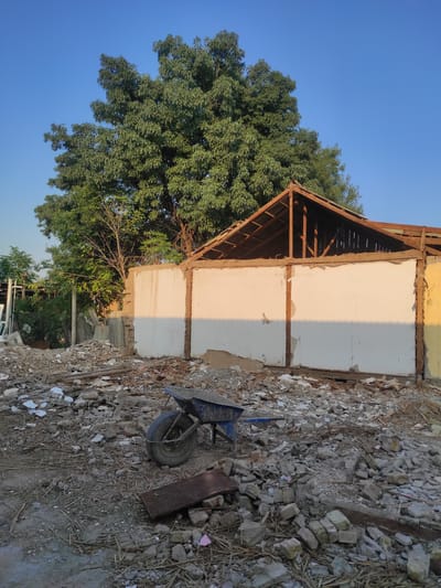 This is a photograph taken outdoors in Gagarin, Uzbekistan, depicting the aftermath of demolition. A partially deconstructed building with exposed wooden roof beams and white walls stands in the background. The foreground is cluttered with rubble, including bricks, concrete, and debris. A blue wheelbarrow with a black tire is positioned among the rubble, suggesting ongoing cleanup or abandonment after demolition. A large, leafy tree is visible behind the building, and the sky is a clear, bright blue. The lighting suggests it is either early morning or late afternoon, casting long shadows.