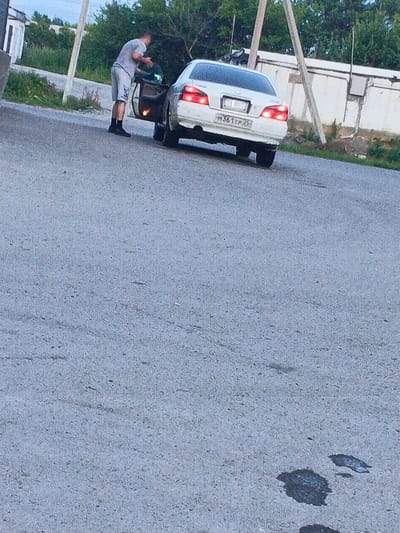 A man wearing a grey t-shirt and shorts, black socks and black shoes is standing next to a white sedan car. The car's driver-side door is open. The man is reaching into the car, possibly putting something inside. The car's license plate is partially visible, showing the characters "M361TP25". The scene appears to be on a paved road, with vegetation and a white wall in the background. The image quality is low, with a grainy texture. The location is identified as Spassk-Dal'nij, Russia.