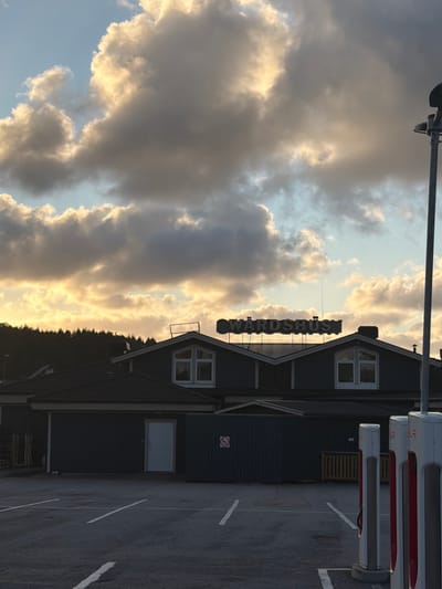 In the city of Hogstorp, Sweden, a parking lot is pictured with several Tesla charging stations in the foreground. In the background, a dark-colored building with a prominent sign reading "VÄRDSHUS" is visible, situated against a backdrop of dark clouds illuminated by the setting sun. A line of trees forms a silhouette behind the building, and a lamppost stands to the right of the frame. The parking lot is marked with white lines.