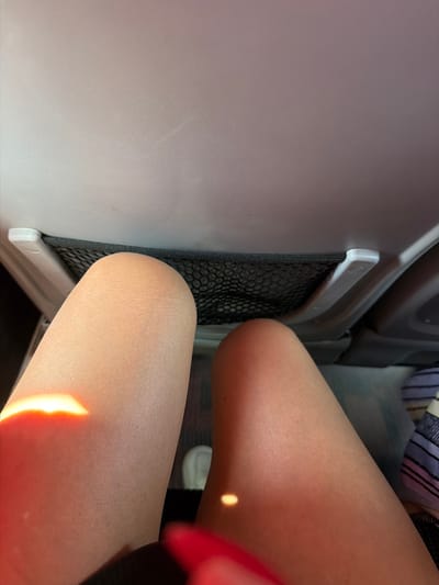 The image shows a close-up view of a person's legs, positioned in what appears to be the seating area of a vehicle. The legs are bare, and one leg is illuminated by a bright, horizontal light source. A black mesh pocket is visible on the back of the seat in front, with white plastic fixtures on either side. The seat material appears to be a light grey or beige. The overall context suggests a travel or transportation setting. The image does not provide any specific visual cues to determine the location within the city of Журавское сельское поселение, Ukraine.
