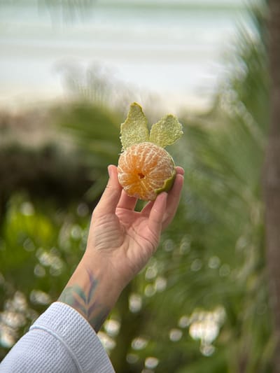 A person's hand, with a tattoo visible on the wrist, holds a partially peeled tangerine. The tangerine has two pieces of peel artfully arranged to resemble bunny ears. The background is softly blurred, showing hints of greenery and a pale, hazy sky, possibly indicating an outdoor setting in Dong Ninh Hoa, Vietnam. The person is wearing a light-colored, textured robe.