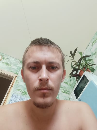 This is a first-person, selfie-style photograph, taken from a low angle, showing the upper body of a light-skinned, fair-haired young adult male. He has short, light brown hair, a thin mustache, and a small goatee. His eyes are open and looking directly at the camera. He is shirtless, with his shoulders and chest visible.
The background appears to be an indoor setting, possibly a bathroom, with patterned wallpaper featuring green leaves and white floral motifs. To the left, a doorway with light brown trim is partially visible. To the right, a white appliance, possibly a microwave or small refrigerator, is present, topped with a potted plant with dark green leaves. The ceiling is a light, neutral color. There is no discernible information in the image to suggest the location in the city of Ушья, Russia.