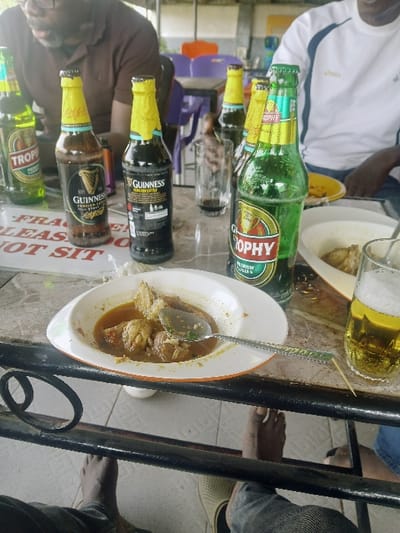 The image captures a casual social gathering around a table, likely in an outdoor or semi-outdoor setting, possibly a patio or a relaxed eatery. The photo is rotated 90 degrees counter-clockwise.

On the dark-colored table, there is an array of food and drinks. Prominently featured are multiple beer bottles and glasses. Two green bottles clearly display "Trophy Lager" on their labels. Two dark brown bottles are visible, identified as "Guinness Foreign Extra Stout." There are also at least two clear drinking glasses; one contains a frothy, amber-colored beverage, presumably beer, while another appears empty.

A white bowl with an orange rim holds a stew or soup, containing pieces of meat, possibly chicken or fish, in a rich, reddish-brown sauce. A spoon is resting inside the bowl. Another white plate, partially visible towards the top right, appears to have additional food, possibly fried chicken pieces. A small wooden toothpick or stick is also seen on the table surface next to one of the glasses.

Several individuals are present, though only parts of them are visible. Towards the top left, a person wearing a white t-shirt with blue trim is seen. In the bottom left, another person wearing a brown shirt, with glasses and a beard, is partially visible. The lower legs and feet of at least two other individuals are discernible beneath the table; one foot is bare, and another is wearing a light-colored shoe.

In the background, colorful plastic chairs (purple, orange) are stacked or placed, indicating a casual dining or seating area. A white sign or sticker, affixed to what appears to be a table leg or a structural support in the bottom center, has partially legible text that reads "FRAGILE" and "PLEASE DO NOT SIT." The lighting suggests it is daytime. The overall atmosphere appears relaxed, with people enjoying food and drinks together.