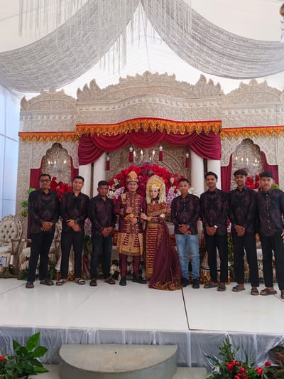 In Meuliek, Indonesia, a wedding ceremony takes place under a white tent adorned with silver drapery and hanging white tassels. A meticulously decorated stage serves as the focal point, featuring ornate, carved white panels with intricate patterns, framed by red velvet curtains and gold trim. Chandeliers hang from the backdrop, illuminating a vibrant display of red and pink flowers.
The central figures are a newly married couple dressed in traditional attire. The bride wears a deep red, intricately embroidered gown with gold accents and a golden headdress. The groom is dressed in a matching deep red and gold outfit, complete with a decorative hat. They stand together, facing forward, possibly posing for a photograph.
Flanking the couple are eight men. The men on the left, from the viewer's perspective, are dressed in matching dark patterned shirts and black pants, some wearing glasses. The men on the right are similarly attired in dark patterned shirts and black pants, except for one who wears blue jeans. All the men are standing in a line, some with hands clasped in front of them. The scene is set on a white stage, with ornate white chairs visible to the left. Green foliage and red flowers are placed at the front of the stage, adding a touch of natural beauty to the celebratory setting.