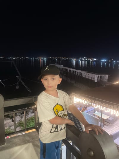 A young boy wearing a white t-shirt with a yellow graphic, blue shorts, and a black baseball cap stands on a balcony at night. He is looking at the camera. Behind him, a body of water is visible, with lights reflecting on the surface from a distant cityscape and a long, white pier with a building. The balcony has a railing and a large, dark circular object in the foreground. The atmosphere is dimly lit with string lights visible on the balcony above and to the right. This scene is consistent with a location in the city of Anapa, Russia, which is known for its coastal setting and waterfront developments.