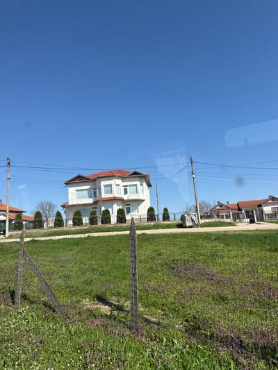 The scene shows a view of residential properties in Poroino, Bulgaria, taken from a vehicle. A two-story, white building with a red roof stands prominently in the foreground, fronted by trimmed evergreen trees and a grassy lawn. Adjacent to the building, a worn wooden fence runs along the edge of a field with mixed green vegetation. Power lines stretch across the blue sky above. Other residential structures with similar roof designs are visible further back in the scene, suggesting a consistent architectural style. The clear sky suggests it is daytime. The focus on residential architecture and green spaces indicates a suburban or rural setting within the city.