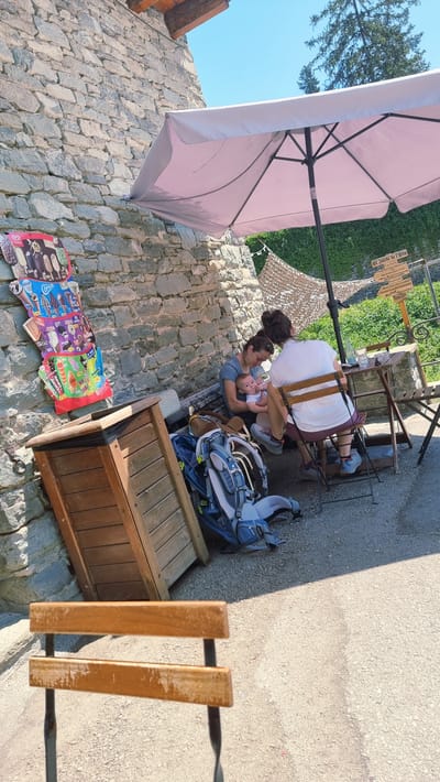 An outdoor scene captures a momentary pause for relaxation and sustenance in Rupit, Spain. In the foreground, an empty wooden chair with a metal frame stands on a gravel-covered ground. To the left, a stone wall forms the backdrop, adjacent to a wooden bin. A colorful display of ice cream advertisements is affixed to the wall above the bin. Under a light pink umbrella, two individuals and a baby are seated at a small table. One person, a woman, is holding the baby, who appears to be an infant, and is feeding it. The other individual is seated opposite them. Two backpacks, one predominantly blue and gray and the other brown, rest on the ground near the seated figures. Beyond the umbrella, a lush green hedge and a signpost are visible. The sky is a clear, bright blue.