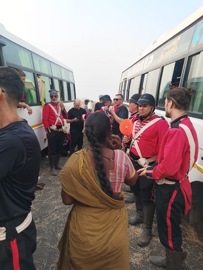 The scene depicts an outdoor gathering of approximately ten individuals, primarily men, situated between two white tour buses parked facing each other in the city of Kottalapalli, India. The ground consists of dark asphalt or dirt scattered with dry, light-colored straw-like debris. The sky is uniformly bright and hazy, indicating either an overcast condition or significant atmospheric haze.
Two large white passenger buses frame the central activity. The bus on the right side of the frame displays "MANIDEEP TOURS & Travels" in yellow and blue lettering on its side. Both buses feature dark tinted windows with light-colored curtains visible inside.
The majority of the individuals are dressed in historical military-style costumes: red long-sleeved coats, black trousers with red vertical stripes down the sides, white cross-body belts, and tall dark boots. Several men also wear black military helmets or shakos.
In the foreground, a woman with dark skin and long, dark hair braided down her back is observed from behind, wearing a multi-colored striped blouse and a gold or tan sari draped over her. She faces a man in a red military uniform and black helmet who holds a small, round, orange fan in his right hand. To the right of this man, another man in a similar uniform, with dark hair tied in a bun and a shaved undercut, stands facing him.
To the left, a man in a black long-sleeved shirt and black trousers with a red stripe is visible from behind. Further left, near the white bus, a man in a red uniform with a patterned bandana on his head and sunglasses is leaning against the bus, with an object near his mouth, possibly smoking. Another man in a black t-shirt is seen in the mid-ground examining a mobile phone. A few other individuals in varying attire are visible further in the background, including a person leaning out of the left bus window. Distant hills are faintly visible under the hazy sky.