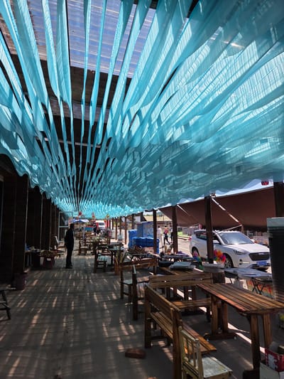 This image captures an outdoor pedestrian area, likely a market or restaurant seating, under a unique light blue fabric canopy. Numerous sheer, light blue fabric strips or sheets are strung overhead, creating a wavy, decorative ceiling that filters sunlight. The sunlight casts intricate, wavy shadows on the ground and wooden surfaces below, indicating a bright, sunny day, possibly morning or late afternoon.

Beneath the canopy, several rustic wooden tables and matching benches are arranged on a paved ground, suggesting a casual dining or resting space. In the background, a white car is parked, bearing a visible license plate "D31613" along with Chinese characters. Near the car, colorful pinwheels add a touch of whimsy. Further back, other structures and what appear to be market stalls or shop fronts are visible. One person, dressed in dark clothing, is seen standing in the mid-ground, their shadow extending across the floor.

A partial sign with Chinese characters "金牌昌..." (Jin Pai Chang...) can be seen on a red object in the background, possibly indicating a brand or business. The setting appears to be a pleasant and functional outdoor area in Jiejinkou, China, offering shade and a place for leisure.