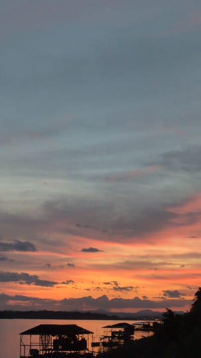 A sunset scene over a body of water in Vientiane Capital, Laos, displays the sky in shades of muted blue, grey, and vibrant orange-pink hues. The clouds are streaked with these colors, creating a dramatic gradient effect. In the foreground, silhouetted against the colorful sky, are several stilted structures, appearing to be small huts or floating platforms, situated on the water. A dark, treed shoreline is visible in the distance, with hints of mountains or hills beyond. The water is calm and reflects the soft colors of the sky. A small dark speck, possibly a bird or an aircraft, is visible in the upper portion of the sky.