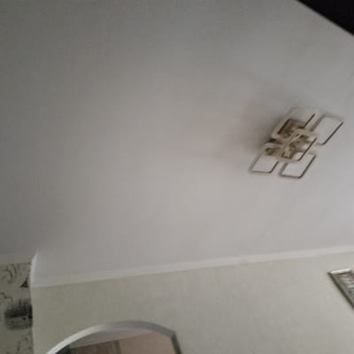 The image shows a ceiling with a modern geometric light fixture. The fixture is made of interconnected rectangular shapes with a metallic finish. The ceiling itself is painted white and appears to be smooth. Below the ceiling, a wall is visible, painted a light, neutral color. To the left of the image, a portion of wallpaper with a decorative pattern is visible. Further down, an arched doorway or opening is partially visible, suggesting an interior space. On the far right, a framed picture is hanging on the wall, though its contents are not discernible. The lighting in the room appears to be ambient, possibly from the fixture itself or other unseen sources. There is no indication of the time of day or weather conditions. The scene depicts an indoor setting, likely a residential room. No people or specific activities are depicted. No text is visible in the image.