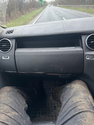 A forensic perspective from the front passenger seat of a vehicle on the A303 road near Wylye, United Kingdom, during active rainfall. The vehicle's black dashboard, featuring circular air vents on both sides and a closed glove compartment, displays dust and light dirt accumulation along its lower portion. In the foreground, the passenger's legs are visible, clad in dark trousers and footwear heavily soiled with dried mud. These rest on a black rubber floor mat also covered in mud. Through the rain-splattered windshield, a wet, two-lane asphalt road extends forward, bordered by green grassy verges and trees. A distant green road sign clearly indicates "A303" and "Wylye" among other destinations. Faint lights of other vehicles are discernible further down the wet road. The scene depicts travel in adverse weather conditions, with the mud on the passenger and floor suggesting recent activity in a muddy outdoor environment.