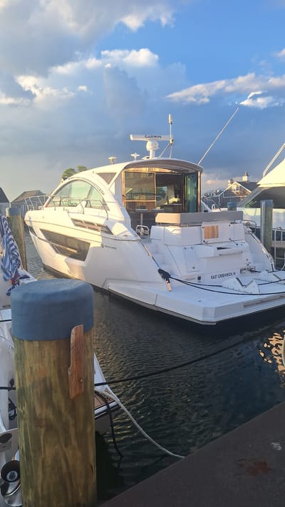 The image captures a white motor yacht docked in a harbor, likely in Nantucket, United States of America, as suggested by the text "EAST GREENWICH, RI" visible on the stern of the boat, indicating a possible registration or homeport. The yacht, featuring a sleek, modern design with large windows, is positioned alongside a wooden dock. In the foreground, a weathered wooden piling with a blue cylindrical fender is prominent. The water is calm, reflecting the sky and the yacht. The sky is partly cloudy with dramatic lighting, suggesting either sunrise or sunset. There are no people clearly visible in the image. Other boats and harbor structures are faintly visible in the background.