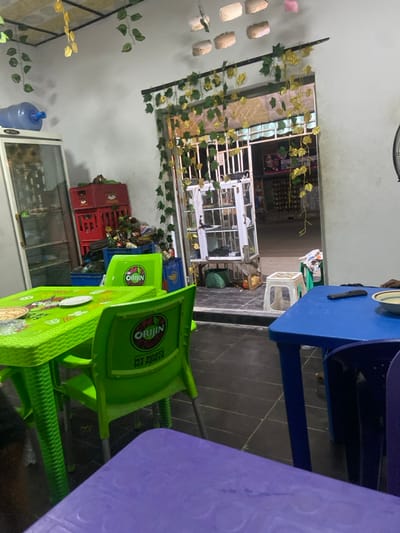 Inside a building, there is a window with leafy decorations. Stacks of crates and boxes with drinks are visible. A green plastic chair, branded with "ORIJIN," stands next to the crates. In the foreground are blue and purple tables with plastic tops. A small white stool is positioned further in, leading into a store-like area.