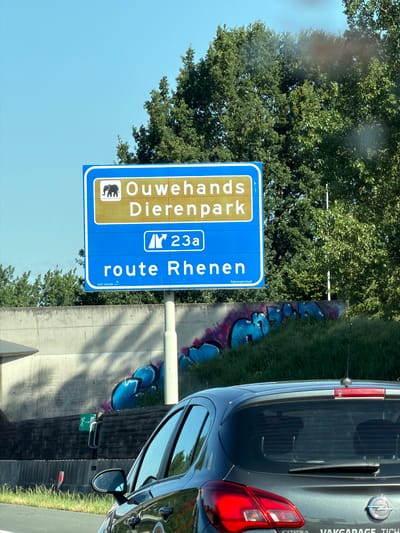 A Dutch road sign for Ouwehands Dierenpark in Rhenen is visible against a bright blue sky and lush green trees. The sign is blue with white text and an elephant graphic. The text reads "Ouwehands Dierenpark" and "route Rhenen." Below the main text, "Rijkswaterstaat" is printed in smaller white letters. The sign is mounted on a white pole. In the foreground, a dark gray car, possibly a hatchback, is driving on a road. Graffiti in blue and purple is visible on a concrete wall behind the sign. To the left of the sign, a green emergency exit sign with a running figure is attached to a metal post. The scene suggests a location in De Klomp, Netherlands, given the reference to Ouwehands Dierenpark.