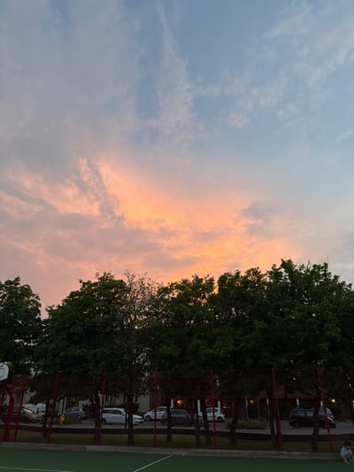 The image depicts a twilight scene in Helsingborg, Sweden, featuring a basketball court in the foreground. The court has green turf with white markings. A red fence surrounds the court, beyond which are trees and parked cars. In the background, there are buildings and more trees. The sky above is a gradient of blue and pink, indicating either sunrise or sunset, with scattered clouds. A lone child is seated on the edge of the basketball court in the lower right corner of the frame. The overall atmosphere is calm and serene.