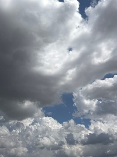 This is a close-up, low-angle shot of a sky filled with dramatic, turbulent clouds. The clouds are predominantly gray and white, with some patches of deep blue sky visible. The lighting suggests it is daytime, with the sun partially obscured by the thick cloud cover, creating a sense of depth and texture. The clouds appear to be moving and forming, with some areas showing bright, illuminated edges, possibly indicating an approaching storm or the aftermath of one. The overall mood is one of grandeur and perhaps a touch of foreboding due to the dark, heavy clouds. The image offers no specific location cues beyond the sky itself, and no text is visible.