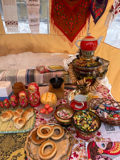 This image captures an outdoor vendor's stall in Plast, Russia, situated under a beige/tan tent with transparent panels revealing a snow-covered ground and distant building in the background, indicating a winter setting.
The scene focuses on a table densely covered with patterned tablecloths, displaying traditional Russian goods and food items. A prominent, ornate gold-colored samovar, painted with red and black floral designs, stands centrally, topped by a red and white teapot. A string of sushki bagels is looped around the samovar's base.
To the left, a graduated set of five red matryoshka nesting dolls with floral patterns is arranged alongside a small wooden barrel and a white rectangular block. A light green rectangular platter holds several small, braided bread rolls. A silver platter presents multiple bubliki bagels.
Other items on the table include a clear glass bowl containing small red apples, a wooden bowl filled with pastel-colored candies, and two additional bowls holding an assortment of individually wrapped, colorful candies. A small black coffee grinder on a wooden base, a red and white teacup with a saucer, a small red box, and a yellow patterned plate are also present. A white paper with a QR code is visible on the bottom right.
Hanging textiles visible in the background include two white embroidered linen pieces and a large red floral shawl with fringe, alongside a darker patterned fabric. No people or active interactions are depicted within the frame.