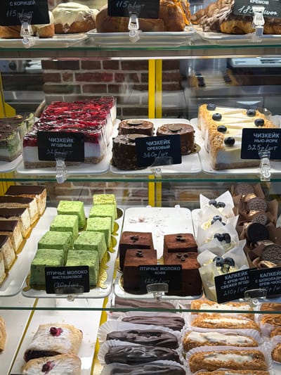 This image captures a brightly lit, multi-tiered retail display case filled with a variety of freshly prepared baked goods and desserts, located in a commercial establishment within посёлок 1-го отделения совхоза "Масловский", Russia.
The display features at least four glass shelves supported by yellow vertical beams. The background exhibits a reflection of a red brick wall. On the top shelf, various sweet rolls are arranged, including cinnamon rolls ("С КОРИЦЕЙ") priced at 115 rubles per piece, and other cream-filled pastries ("КРЕМОМ").
The second shelf from the top presents three distinct cheesecake varieties: a large rectangular "ЧИЗКЕЙК ЯГОДНЫЙ" (Berry Cheesecake) with a vibrant red berry topping, priced at 325 rubles per piece; several smaller, individual "ЧИЗКЕЙК ОРЕХОВЫЙ" (Nut Cheesecake) with dark chocolate coating and golden embellishments, priced at 360 rubles per piece; and a large rectangular "ЧИЗКЕЙК НЬЮ-ЙОРК" (New York Cheesecake) garnished with blueberries, priced at 325 rubles per piece. All items are presented on white rectangular plates.
The third shelf features a selection of layered cakes and individual desserts. On the left are slices of layered cakes with dark tops. Adjacent to them are rows of small, rectangular "ПТИЧЬЕ МОЛОКО ФИСТАШКОВОЕ" (Ptyche Moloko Pistachio) cakes, colored green and priced at 130 rubles per piece. To their right are similar "ПТИЧЬЕ МОЛОКО КОФЕ" (Ptyche Moloko Coffee) cakes, dark brown with two coffee beans on top, priced at 125 rubles per piece. Further right, white cupcakes topped with cream and blueberries ("КАПКЕЙК С КРЕМ-ЧИЗ СВЕЖЕЙ ЯГОД") are displayed in paper wrappers, priced at 210 rubles per piece, alongside other cupcakes topped with Oreo cookies ("КАПКЕЙК С ШОКОЛАДНОЙ КРЕМ-ЧИЗ").
The bottom shelf showcases pastries with fruit filling, powdered sugar, and two types of éclairs: one row of dark chocolate-coated éclairs and another row of lighter-colored, cream-filled éclairs. Each item or group of items is accompanied by a black price tag with white Russian text and a price in rubles, affixed with clear plastic clips. No individuals are visible in the frame.