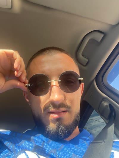 A close-up, angled upward shot taken from inside a vehicle shows a person holding up round, rimless sunglasses. The vehicle interior is visible, including the headliner, a passenger assist handle, and a sunroof. The person has a shaved head and a beard. A blue and white polka-dot shirt is partially visible. The specific location, Novo Leski, Bulgaria, is not discernable from the visual information.