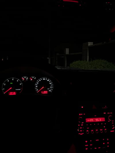 The image shows the interior of a car at night. The dashboard is illuminated with red lights, displaying the car's speed, RPM, fuel level, and temperature. The car's radio is also visible, with the display showing "FM 93.3". The exterior environment is dark, with some greenery and a fence visible through the windshield. It is impossible to determine the specific location in the city of Ponebel, Ukraine, from the provided image.