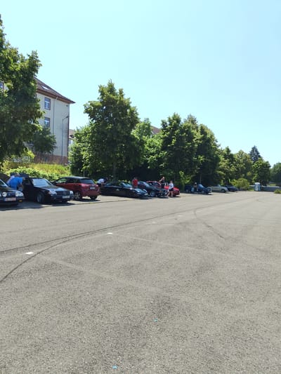 A group of people are gathered around several parked cars in a lot. The cars appear to be older models and include a black Mercedes-Benz, a red SUV, and a black convertible. Some of the people are standing and talking, while others are leaning into the cars, possibly examining them. The scene is set against a backdrop of trees and a building. The sky is clear and blue. The location is the parking lot of the former Elsa-Brandström-Kaserne in Pirmasens, Germany.