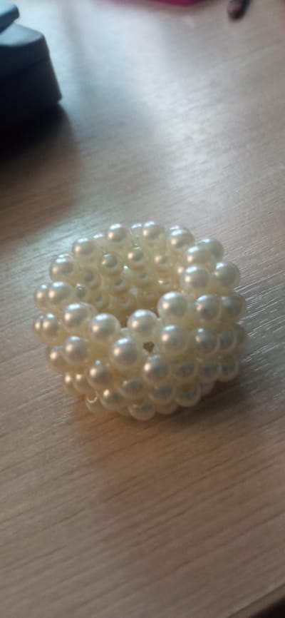 A round, textured object made of numerous small, iridescent white beads is positioned on a light brown, wooden surface. The object is centrally located in the frame and appears to be a decorative item, possibly a bracelet or a hair accessory. The surface beneath it has visible wood grain and a slightly reflective quality, indicating it is likely a table or desk. In the upper left portion of the image, a dark, out-of-focus object is partially visible, its shape suggesting it could be a piece of electronic equipment. The lighting suggests an indoor environment, with a soft light source casting subtle shadows. There are no people or animals present in the image. The specific location of Tatishchevo, Russia, is not visually discernible from the image itself.