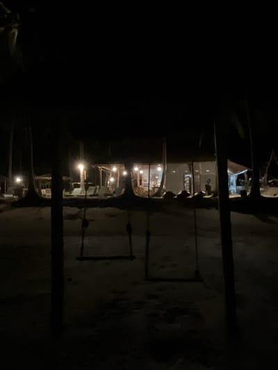 This is a nighttime photograph taken at a beach resort in Legonipah, Indonesia. In the foreground, there are two empty swings hanging from unseen structures, casting dark shadows on the sandy ground. Beyond the swings, illuminated by artificial lights, is a row of buildings that appear to be part of the resort. These buildings have open-air sections and are lit from within, revealing glimpses of furniture and possibly patrons. Palm trees are visible on either side of the scene, their silhouettes dark against the night sky. Lounge chairs are also visible in front of the resort buildings, suggesting a relaxation area. The overall atmosphere is tranquil and dimly lit.