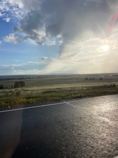The image depicts a vast, open landscape in Kipchak-Askarovo, Russia, with a prominent road running vertically along the right side. The road appears wet, possibly from recent rain, reflecting the overcast sky. The left side of the image is dominated by a dramatic sky filled with clouds. The sun is visible, partially obscured by clouds, casting rays of light that create a dramatic effect. The landscape itself consists of fields and sparse vegetation, with a few scattered trees visible in the distance. The overall impression is one of a rural or semi-rural setting. The time of day is difficult to determine precisely due to the cloud cover and the angle of the sun, but the lighting suggests it could be late afternoon or early evening. The weather appears to be unsettled, with a mix of clouds and sunshine. There are no people or specific objects that stand out, and no visible text.