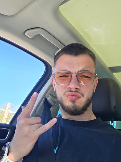 A man with a beard and tinted glasses is in a car with a sunroof on a sunny day. He is wearing a black shirt and giving a peace sign with his right hand. The location is not discernible from the image.
