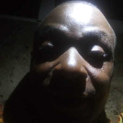 A close-up, frontal image depicts the upper facial features of an adult individual in the city of Ibreda, Nigeria. The subject, presenting dark skin pigmentation, is illuminated by a strong, overhead light source, creating pronounced specular highlights across the forehead, nose, and upper cheeks. Deep shadows obscure the eyes, lower face, and the surrounding environment, which appears uniformly dark. The individual's gaze is directed towards the camera. No discernible objects or further environmental details are visible.