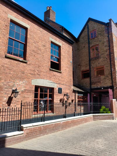 The image shows a courtyard area in front of a brick building with multiple windows and a chimney. The building has red brick walls and red-framed windows, some of which are divided into multiple panes. There is a black metal fence running along the front of the building, separating it from a paved courtyard. The sky is clear and blue. The building appears to be a historic structure, possibly a pub or restaurant, given the style of the windows and the presence of outdoor seating areas visible through the windows. The context provided suggests this location is in Ludlow, United Kingdom.