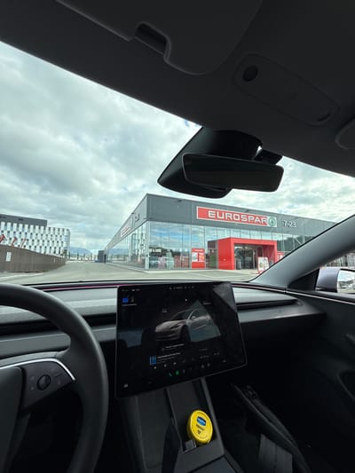 The image shows the interior of a Tesla vehicle parked in front of a Eurospar supermarket located in Harstad, Norway. The Eurospar store has a large red entrance and is identifiable by its signage. Adjacent to the Eurospar is another modern building with a patterned facade. The sky is overcast, and mountains are visible in the distance. Inside the car, the steering wheel and the central touchscreen display are visible. The touchscreen shows what appears to be music playback, with the song title "Unbreakable" by Sam Gray displayed. A yellow container of Vaseline is placed in the center console. The overall scene depicts a mundane moment of a car parked in a commercial area on a cloudy day.
