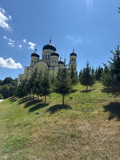 The image depicts a bright, sunny day at the Hancu Monastery in the village of Ciuciuleni, Moldova. The primary subject is the monastery itself, a large, yellow-colored building with multiple black domes, each topped with a cross. The monastery is situated on a gentle, grassy slope. In the foreground, a line of small evergreen trees is planted, casting shadows on the lawn. The sky is a vibrant blue with scattered white clouds. The overall scene is serene and picturesque, showcasing religious architecture within a natural, well-maintained environment.