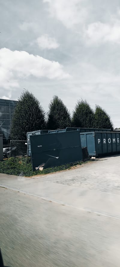 This media file captures an outdoor scene in Fellbach, Germany. The primary objects are several large, dark green industrial dumpsters or roll-off containers positioned in a line. One dumpster clearly displays white lettering spelling out "PROME" on its side. Behind the dumpsters, a row of lush, dark green trees stands tall, some appearing to be manicured. Further in the background, a modern building with a glass facade is visible, reflecting the sky. A chain-link fence runs in front of the trees and the building. The ground in the foreground is a paved area, possibly a parking lot or road, with a grassy strip between it and the dumpsters. The sky is overcast with soft, white clouds, suggesting a daytime scene with diffused light, perhaps mid-morning or afternoon. The overall mood is neutral and industrial. There is no visible activity taking place, implying the image was captured as a static observation. The slight blur in the foreground, particularly on the paved area, suggests the photo may have been taken from a moving vehicle.