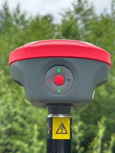 A close-up, eye-level, medium shot features the top portion of a grey and red surveying device mounted on a black carbon fiber pole. The device has a red circular button in the center, surrounded by an array of icons. To the left of the button, a red circle with a horizontal line inside indicates a power button. Above the red button, a green Bluetooth symbol is illuminated. Below the red button, two green arrows point up and down, suggesting a leveling function. To the right of the red button, a grey icon resembling brackets with a central dot is present. A yellow sticker with a black lightning bolt inside a triangle is affixed to the pole directly below the device. The background is heavily blurred, consisting of green foliage under an overcast sky. The scene appears to be outdoors, likely in a natural or semi-rural setting. The city of Murtygit, Russia, is not discernible in the image.