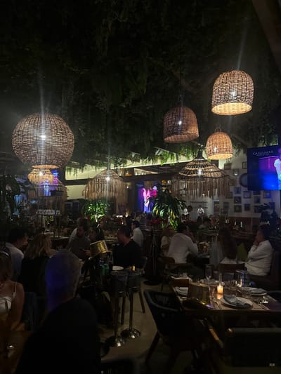 An outdoor restaurant at night with multiple tables of people dining. Wicker lantern-style lights hang above, illuminating the scene. Lush greenery surrounds the dining area. A screen displaying content hangs behind the dining area. The overall ambiance is lively and intimate.