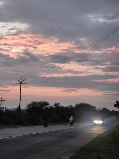 A van and two motorcycles are on a road in Dattapur, India, as the sun sets. The sky is filled with pink and gray clouds. The van's headlights are on, illuminating the road and creating a hazy glow. A person is riding a motorcycle in front of the van, and another motorcycle is visible further down the road. Trees and power lines line the road. The road appears wet, suggesting recent rain.