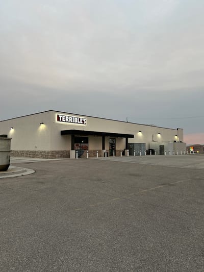 A side view of a white building with a dark awning and a prominent illuminated sign that reads "TERRIBLE'S". The building has several entrances and windows, and is lit by outdoor lights. The foreground is an asphalt parking lot. In the background, distant mountains are visible under a cloudy sky with hints of a sunset. The scene appears to be in a commercial area. There are no people visible in the image. The time of day is likely dusk.