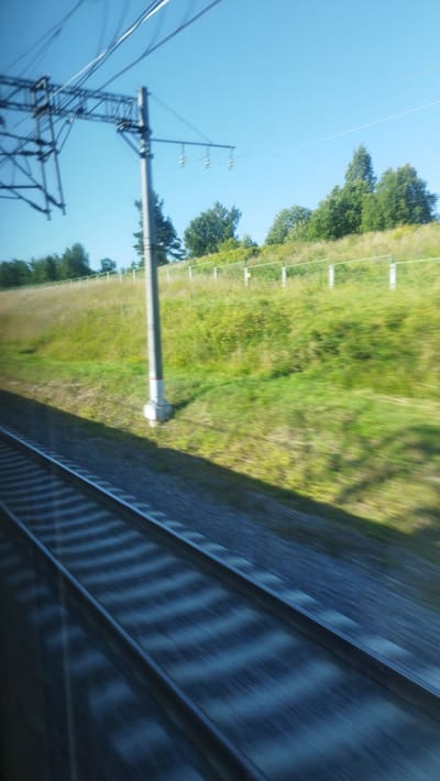 This image captures a view from a moving train in Pashino, Russia. The primary subjects are the train tracks stretching into the distance, the green, grassy landscape, and the blue sky above. Overhead, power lines and a support structure are visible, indicating the train is on an electrified line. The landscape is characterized by a gently sloping hill covered in lush green vegetation and scattered trees. A fence runs along the top of the hill. The scene is bathed in sunlight, suggesting it is daytime, likely late morning or afternoon given the clear, bright sky. There is a sense of movement due to the motion blur, particularly noticeable in the train tracks and the surrounding scenery. No people are visible in the frame. The overall impression is one of travel through a rural or semi-rural area under fair weather conditions.