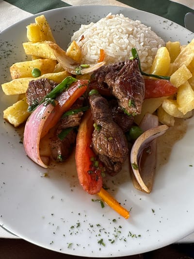 The image shows a close-up, overhead view of a plate of Lomo Saltado, a Peruvian dish, likely served in a restaurant in Lurín, Peru. The dish consists of stir-fried beef, french fries, rice, red onion, tomatoes, and green onions, garnished with chopped parsley. The food is arranged on a white plate, which is placed on a surface covered by a green and white checkered tablecloth. The lighting suggests an indoor setting, possibly during daylight hours. There are no people visible in the image, and the focus is entirely on the food.