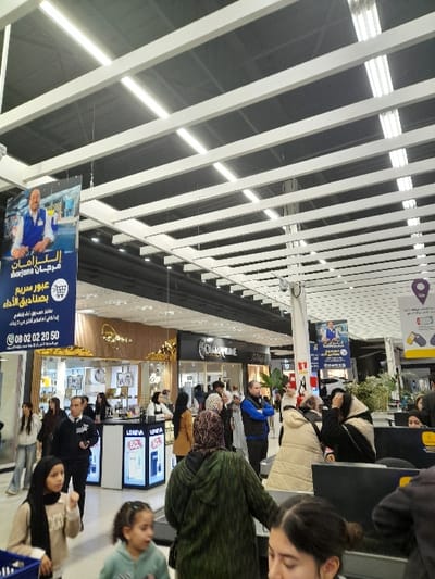 The image shows an indoor scene, likely a shopping mall or airport terminal, with a high ceiling featuring parallel white beams. A variety of shops and people are visible in the background. Prominent signage includes an advertisement for "Marjane," an establishment offering express checkout services. Text in Arabic is present on the signage, indicating a region where Arabic is spoken. The scene suggests a busy commercial environment.
