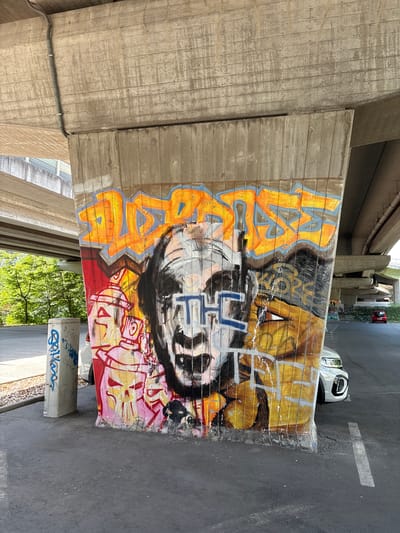 The image depicts a concrete pillar underneath a highway overpass in Erlangen, Germany. The pillar is covered in graffiti, featuring a large, distorted face with exaggerated features, an orange and blue abstract design above it, and spray-painted cans and a skull-like figure to the left. The graffiti extends around the sides of the pillar. To the left of the pillar, a smaller concrete post also has graffiti. A white car is parked to the right of the pillar, and a red car is visible further in the background. Trees and other parts of the overpass structure are visible in the background. The ground is asphalt.