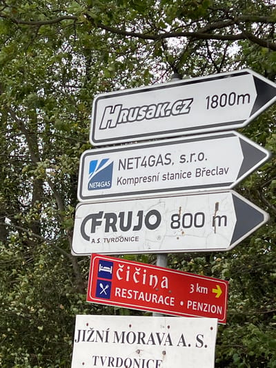 This media file captures a cluster of directional signs against a backdrop of lush green trees and foliage, suggesting an outdoor setting, possibly a park or a natural area. The signs are oriented at an angle, and the foliage partially obscures them, indicating a natural, perhaps slightly overgrown, environment. The lighting suggests it is daytime, and the overall atmosphere is casual and informative.

The signs themselves are the primary subjects. They are a mix of shapes and colors, primarily white with black text, and one red sign with white and yellow text. The visible text on the signs indicates various destinations and distances. One sign reads "HRUSAK.CZ 1800m," and another "NET4GAS, s.r.o. Kompresní stanice Břeclav." A third sign states "FRUJO 800 m" with "A.S. TVRDONICE" below it. A prominent red sign displays "čičina" with a yellow arrow pointing right, followed by "3 km → RESTAURACE • PENZION." Another white sign reads "JIŽNÍ MORAVA A. S." with "TVRDONICE" beneath it. The presence of business names like "NET4GAS," "HRUSAK.CZ," and "FRUJO," along with indicators for "RESTAURACE • PENZION," suggests this is a tourist or travel-oriented location, likely in or near Hrušky, Czechia, as indicated by the location context. There are no people visible in the image. The signs are functional objects providing navigational information. The weather appears to be pleasant enough for outdoor activities, given the clear visibility and the vibrant green of the trees.