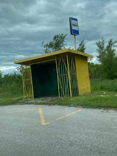 An exterior, eye-level, slightly tilted shot shows a yellow, open-air bus stop structure. The structure has a flat roof and is supported by yellow metal bars that also serve as a partial enclosure on the sides. The back wall of the bus stop is painted dark green. A bus stop sign with a white bus silhouette on a blue background is mounted on a thin pole to the right of the bus stop. The bus stop is situated next to a grey asphalt road with yellow painted markings, likely indicating a parking space or directional lines. Green grass and various trees are visible behind the bus stop, and the sky above is overcast with dark grey clouds. Litter is scattered on the grass. The scene is located in the village of Nadezhdino, Russia. There are no people present in the image.