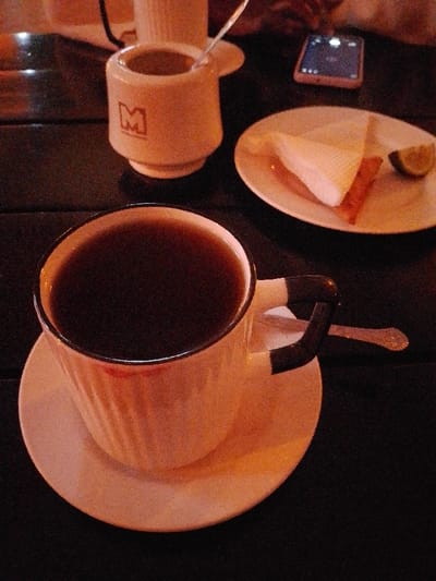 A table displays two cups of coffee, one larger than the other, and a small plate holding a slice of cake with a lime wedge. A smartphone sits near the plate. A spoon rests behind the larger coffee cup. The lighting is warm and dim, creating a cozy atmosphere. A faint lipstick mark is visible on the rim of the larger cup.