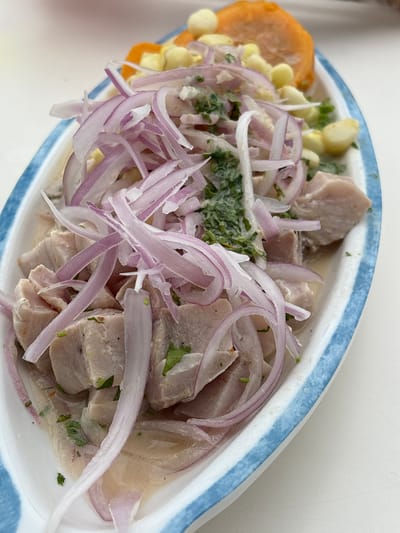 A close-up, overhead shot shows a dish of ceviche served in an oval white dish with a blue trim. The ceviche consists of chunks of white fish, sliced red onions, corn kernels, and a few pieces of sweet potato. Chopped cilantro is scattered throughout the dish. The ceviche is in a light-colored liquid, presumably lime juice. The dish is placed on a white surface, likely a table. The background is blurred, but appears to be an outdoor setting, possibly a restaurant or cafe in La Punta, Peru.