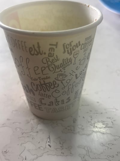 A light-toned, disposable paper cup is positioned upright on a flat, light-colored surface in the city of Lehvaz, Armenia. The cup's exterior is adorned with grey text, including words such as "COFFEE," "est.," "TEA," "for you," "Best Quality," "Fair Trade," "Latte," "Cakes," "TASET," "ROAST," and "Love," alongside small illustrations of a coffee cup, a wine glass, and a heart. Minimal brown residue is visible along the interior rim of the cup. The surrounding surface is marked by numerous irregular, dried liquid stains, appearing light brown to greyish in color, extending outwards from the cup's base. A blurred blue object is partially discernible in the upper right portion of the background. No individuals are present within the frame.