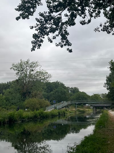 This is a photograph of the Canal de l'Ourcq in Claye-Souilly, France, on a cloudy day. In the foreground, the still water of the canal reflects the surrounding greenery and sky. On the left bank, lush vegetation lines the water's edge. A figure, appearing to be a person, is visible in the distance on the left bank, engaged in an activity near the water. Further along the canal, a pedestrian bridge crosses the water, with another bridge visible beyond it. The bridge has railings and a pathway, and a few people are walking on it. The background is filled with dense trees and foliage. Overhead, power lines are strung across the sky, partially obscured by the branches of a tree in the upper portion of the frame. The overall scene depicts a tranquil, natural setting within an urban or semi-urban environment.
